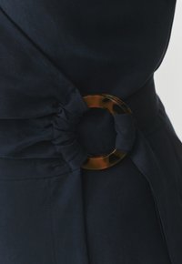 Dettaglio di un abito in tessuto blu navy con materiale arricciato in vita e un'accattivante fibbia circolare in tartaruga. Texture complessiva liscia.