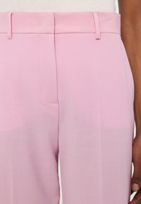 A világos rózsaszín nadrág sima textúrával, fazonra szabott derékrésszel és rejtett oldalsó zsebbel rendelkezik, amely a minimalista dizájnt hangsúlyozza.