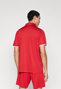 Κόκκινο αθλητικό πόλο μπλουζάκι με γιακά και λευκή διακοσμητική λεπτομέρεια, συνδυασμένο με κόκκινα σορτς. Ομαλή υφή, σχεδιασμένο για άνεση και δραστήρια χρήση.