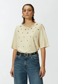 Bež majica kratkih rukava s dekorativnim čavlima i šljokicama, zaobljenog izreza s metalnim detaljima, u kombinaciji s trapericama visokog struka u plavoj boji i crnim remenom.