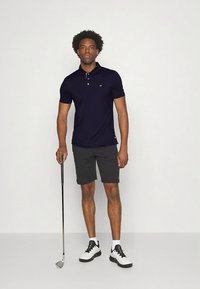 Μπλε ναυτικό πόλο με κοντά μανίκια και γιακά με τρία κουμπιά, συνδυασμένο με γκρι σορτς. Το σύνολο ολοκληρώνουν μαύρα και λευκά παπούτσια για γκολφ.