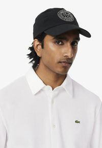 Schwarze Kappe mit gesticktem Logo, weißes Poloshirt mit Knopfverschluss und strukturiertem Stoff, mit kleinem grünen Krokodil-Emblem.