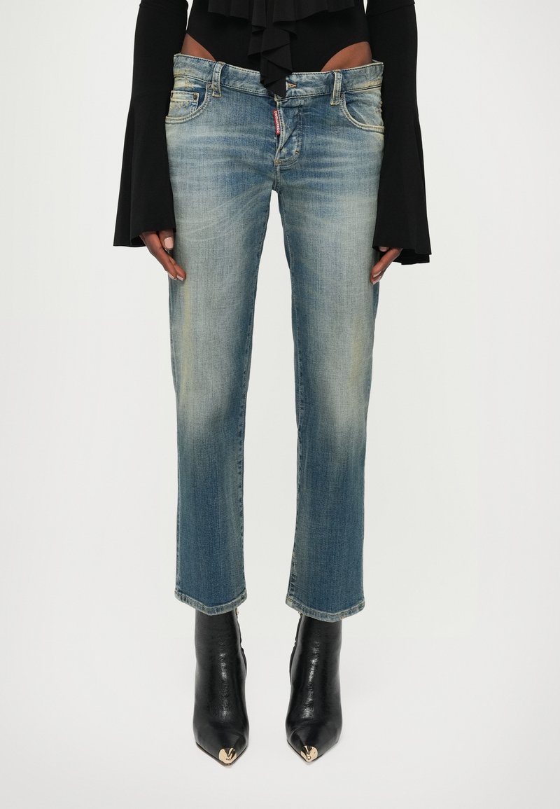 Άτομο που φοράει ξεθωριασμένο μπλε τζιν παντελόνι με ίσια γραμμή, μαύρο μακρυμάνικο τοπ με φαρδιές μανσέτες και μαύρα μποτάκια με μυτερή μύτη και χρυσές λεπτομέρειες.