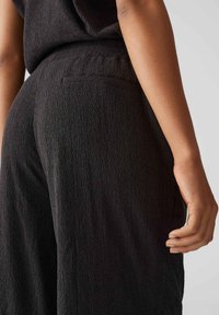 Pantalon noir texturé avec une poche arrondie discrète à l'arrière, présentant un tissu doux et légèrement froissé, ainsi qu'une coupe décontractée au niveau de la taille.