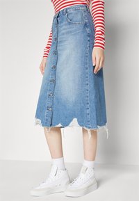 Denim A-linijska krila v svetlo modri barvi z razfrkljanim valovitimi robovi, z gumbi na sprednji strani in dolžino do kolen. V kombinaciji z belimi tenisicami.
