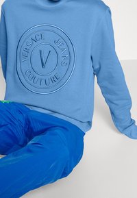Personne portant un sweat-shirt bleu clair avec un logo en relief de Versace Jeans Couture et un pantalon bleu vif, assise sur un fond blanc.