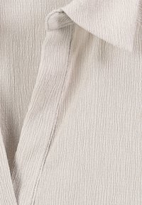 Nahaufnahme von leicht beige strukturiertem Stoff mit einem zerknitterten Muster, das sanfte Falten und ein Kragendetail zeigt.