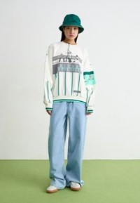 Λευκή μπλούζα με πράσινες λωρίδες και γραφικό τύπωμα, συνδυασμένη με ανοιχτό μπλε παντελόνι σε φαρδιά γραμμή και λευκά αθλητικά παπούτσια. Επίσης, ένα πράσινο καπέλο τύπου bucket.