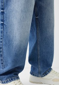 Ljusblå, blanka jeans med lös passform och synliga sömnadsdetaljer, tillsammans med vita sportskor.