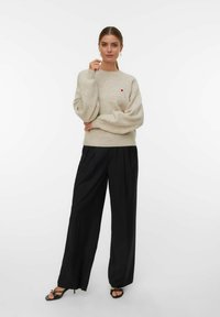 Donna che indossa un maglione beige con un piccolo cuore rosso, pantaloni neri a gamba larga e sandali neri con tacco, in piedi con una mano vicino al viso.