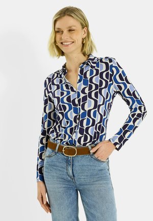 Camisa de manga larga con botones en patrones geométricos azul y navy, combinada con jeans azul claro y cinturón marrón. Tejido liso y ligero.