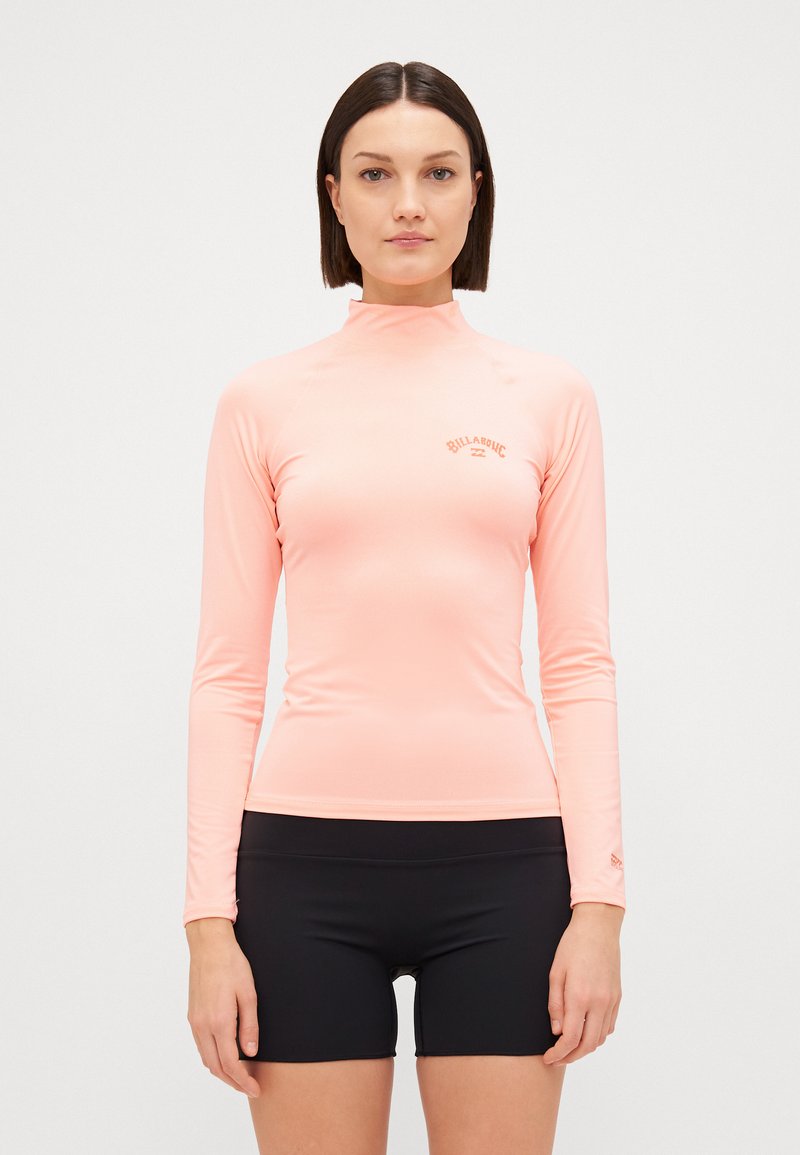 Rash guard a maniche lunghe in morbido materiale rosa chiaro. Presenta un collo alto e un piccolo logo sul petto, abbinato a pantaloni corti neri. Texture liscia.