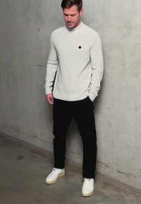 Uomo con maglione grigio chiaro, pantaloni neri e sneakers bianche, in piedi contro un muro di cemento, guarda verso il basso con una mano in tasca.