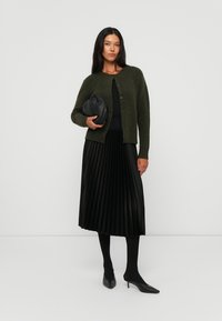 Dark green knit cardigan, pleated black midi skirt, black stiletto heels, and a cinched black handbag. Simple, textured details throughout.