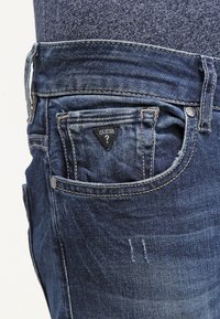 Vaqueros de denim en azul oscuro con una textura desgastada, bolsillos frontales y un parche triangular negro con el logo 'Guess' en el bolsillo derecho.