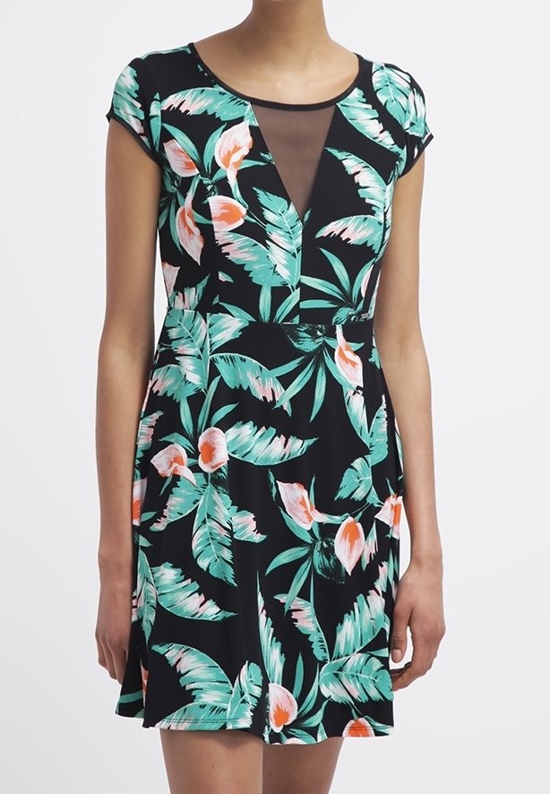 Femme portant une robe noire à manches courtes avec des feuilles tropicales vertes, des fleurs orange et un insert en V transparent.