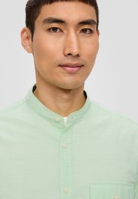 Hele roheline nööpidega shirt, millel on seisva kaelusega, peen triibuline tekstuur ja rinna-tasku. Nööpide värv on sarnane.