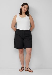 Schwarze Baumwollshorts mit umgeschlagenen Säumen, elastischem Bund, kombiniert mit einem weißen Tanktop und braunen Flip-Flops, vor einem neutralen Hintergrund.