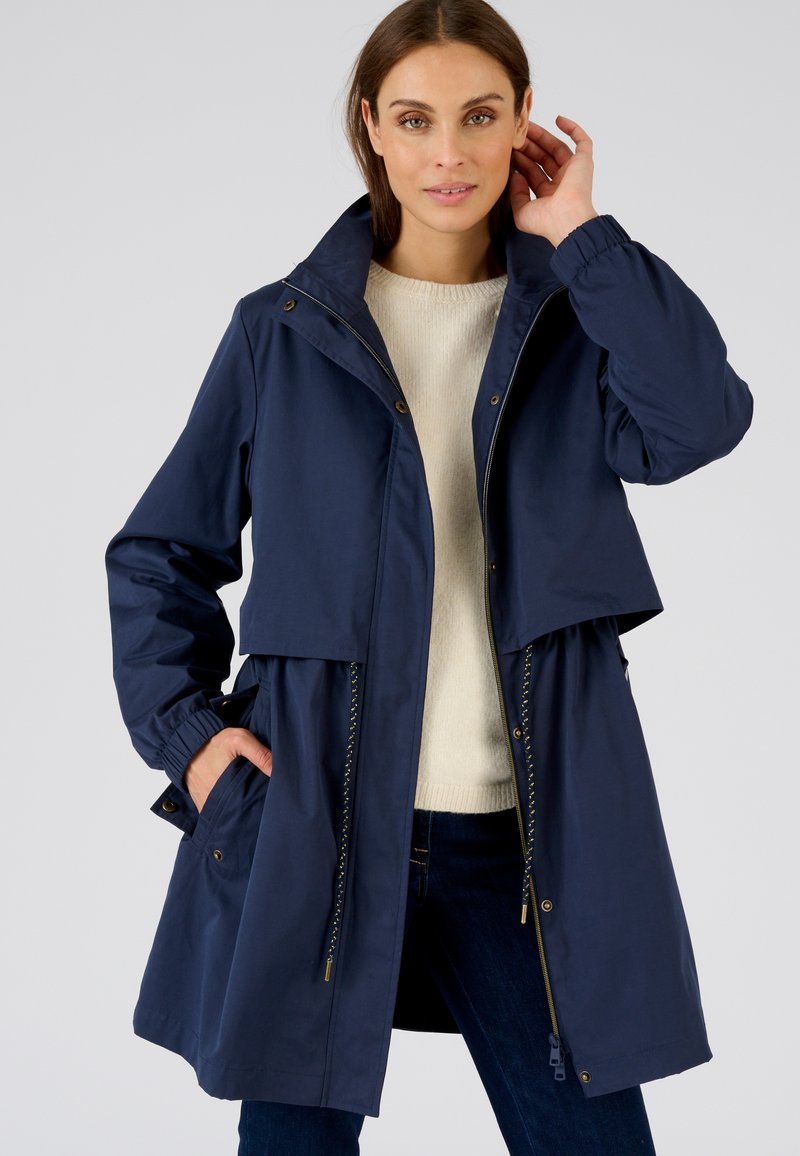 Veste imperméable bleu marine avec un col haut, deux poches avant, des poignets élastiques et un design stratifié. Le matériau semble lisse et durable.