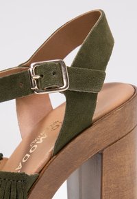 Bretelle de cheville en daim vert avec boucle dorée sur une sandale à talon haut en bois, montrant la semelle intérieure en cuir tanné rembourrée et les détails de couture.