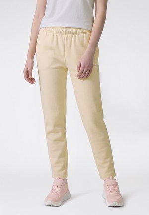 Femme portant un pantalon de survêtement jaune clair avec taille élastique et des baskets roses, debout devant un fond blanc.