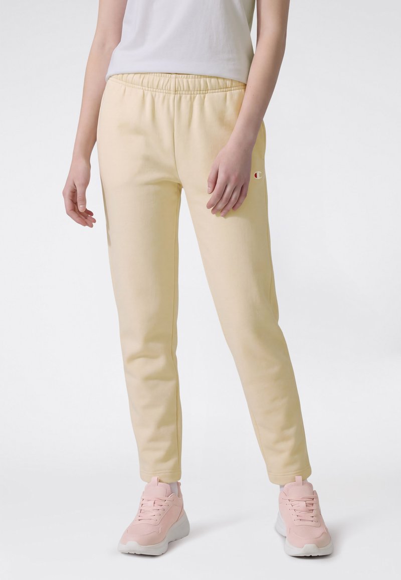 Femme portant un pantalon de survêtement jaune clair avec taille élastique et des baskets roses, debout devant un fond blanc.