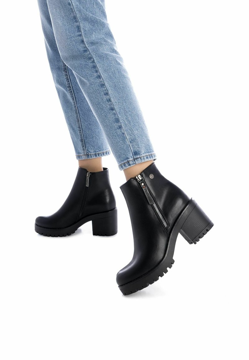 Botas de cuero negras hasta el tobillo con un tacón grueso y suela de goma con relieve. Detalle de cremallera lateral y textura suave. Combinadas con jeans de color azul claro.