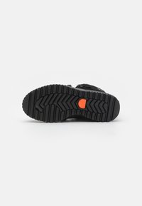 Suela de zapato negra con un patrón texturizado en zigzag; presenta un logotipo de acento naranja. Material de goma para tracción y durabilidad.