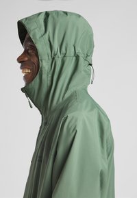 Grüne, wasserdichte Jacke mit Kapuze, die eine glatte, glänzende Textur und Kordeln hat. Frontreißverschluss und Logo auf der Brust sichtbar.