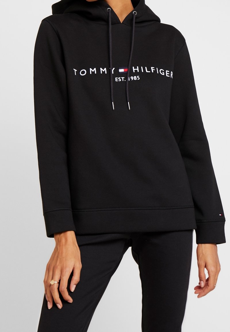 Sweat à capuche noir avec le texte "Tommy Hilfiger EST. 1985" et un logo sur la poitrine, porté par une personne portant un pantalon noir et une bague à la main gauche.