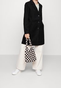 Cappotto nero lungo con due tasche anteriori e bottoni, abbinato a pantaloni beige larghi e sneakers bianche. Tiene una borsa a quadri nera e crema.