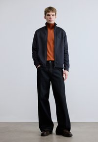 Σκούρο ναυτικό μπουφάν με ψηλό γιακά, πορτοκαλί ζιβάγκο και φαρδιά σκούρα τζιν παντελόνια. Καφέ παπούτσια ολοκληρώνουν την καθημερινή εμφάνιση.