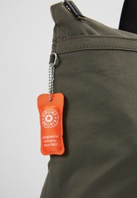 Sac en tissu vert olive avec une étiquette de la marque Kipling orange suspendue à une chaîne à billes argentée près du coin supérieur.