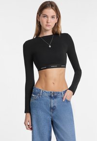 Top corto negro de manga larga con banda con logo. Combinado con jeans de mezclilla azul claro de tiro alto. Textura suave, diseño ajustado.