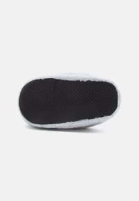 Valkoinen pörröinen slipperin pohja, jossa on karhea musta kumipohja, joka sisältää pieniä pisteitä pitoa varten. Pyöreä muoto, jossa on pehmeä, pörröinen reuna.