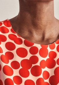 Röd prickig mönster på en ljus bakgrund, tillverkad av len tyg med rundad halsringning. Inga ytterligare detaljer eller accenter.