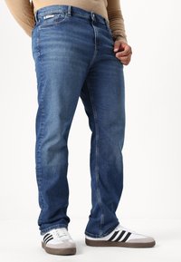 Vaqueros de denim azul con un diseño de pierna recta, con una cintura de altura media y efectos de desgaste sutiles. Combinados con deportivas blancas y acentos grises.
