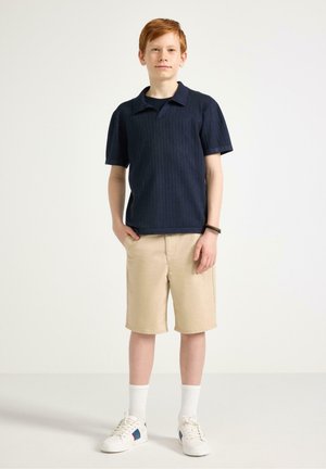 Dreng med rødt hår står med hænderne i lommerne, iført navyblå kortærmet skjorte, beige shorts, hvide sokker og hvide sneakers på hvid baggrund.