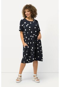 Navyblaues Kleid mit großen weißen Punkten, kurzen Ärmeln, weit geschnitten und mit Taschen. Hergestellt aus weichem Stoff, fällt bis zur Mitte der Wade und wird mit klobigen weißen Sandalen kombiniert.