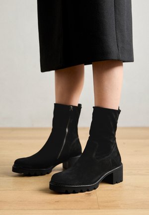 Femme portant des bottes en daim noires mi-mollet avec talons carrés et fermetures éclair sur les côtés, associées à une jupe noire longueur genou, debout sur un sol en bois.