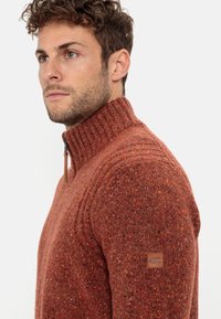 Strukturierter, orange-roter Pullover mit hohem Kragen, Reißverschluss und subtilen Farbtupfern. Besitzt ein Logo-Patch auf der linken Schulter.