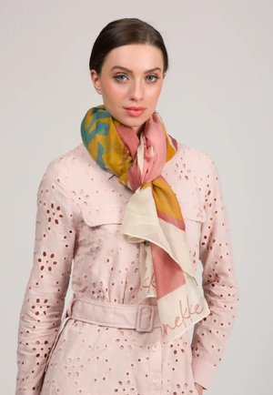 Mujer con una chaqueta rosa de ojales con cinturón y una bufanda multicolor atada al cuello, posando frente a un fondo liso.