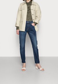 Ljus beige, oversized jacka med fyra fickor över en mörkgrön skjorta, parat med blå smala jeans och vita vävda klacksandaler.