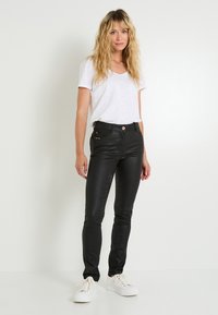 Pantalones ajustados de piel sintética negra con bolsillos laterales, adornados con tachuelas doradas, combinados con una camiseta blanca de cuello en V y zapatillas blancas.