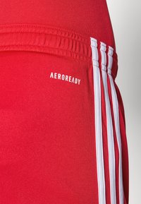 Röda atletiska shorts gjorda av stretchigt tyg med en slät yta, med tre vita ränder på sidan och "AEROREADY"-logotyp.
