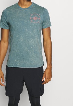 Homme portant un t-shirt à manches courtes bleu délavé avec un petit logo circulaire rose et un short noir, sur fond blanc uni.