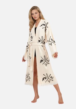 Donna bionda che indossa una lunga veste crema con simboli astratti neri, legata in vita, in piedi a piedi nudi con una gamba leggermente avanti.