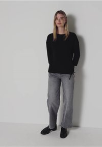 Schwarzes Langarmoberteil, lockere Passform, kombiniert mit grauen weit geschnittenen Jeans. Das Modell trägt schwarze Stiefeletten. Einfache, einfarbige Farben und minimales Design.