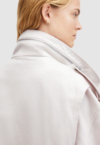 Chaqueta gris claro con un cuello alto, que presenta una cremallera visible y una textura suave. El diseño es minimalista, sin patrones.