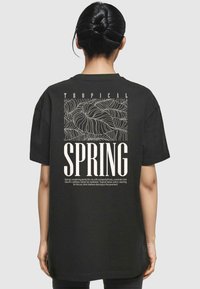 Sort bomulds T-shirt med et stort grafisk print på ryggen med teksten "TROPICAL SPRING" og detaljeret linjekunst af tropiske blade.